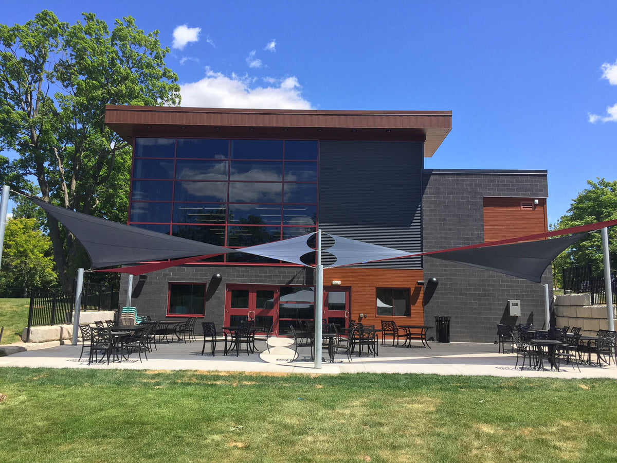 Multiple overlapping shade sails installed at varying heights in front of a modern commercial building, demonstrating proper slope design for effective rainwater drainage and enhanced wind stability in a high-use outdoor seating area.